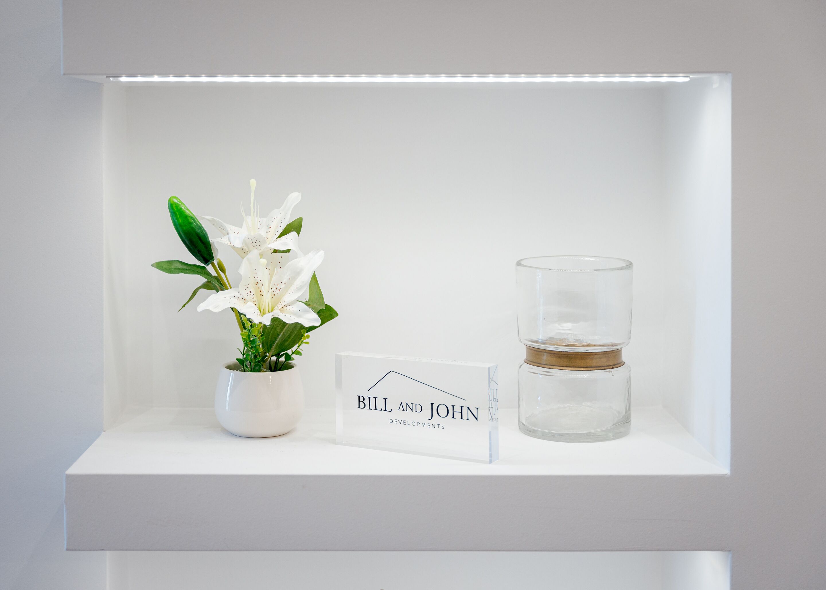A white shelf with a potted white lily, a clear glass candle, and a sign reading "BILL AND JOHN DEVELOPMENTS".