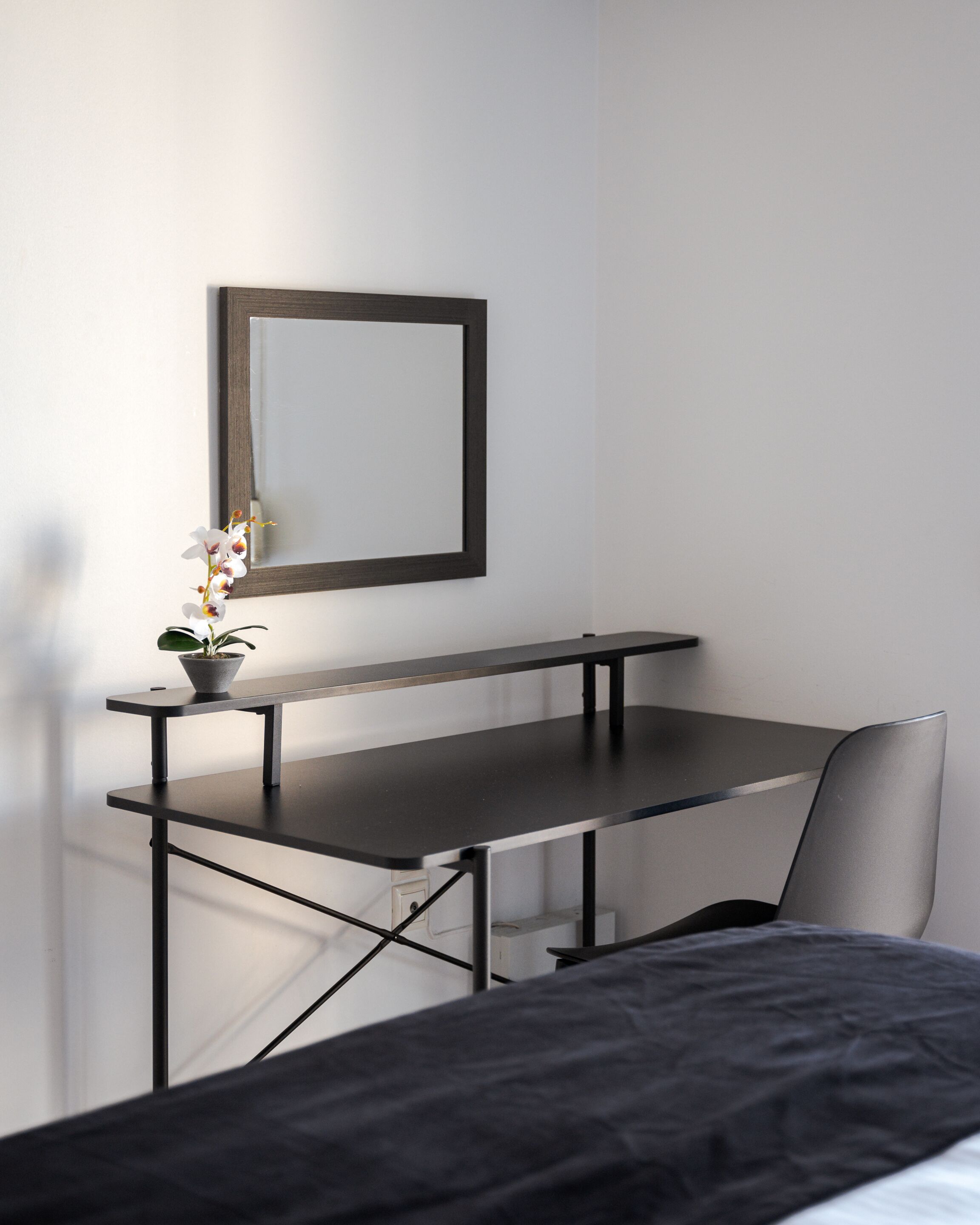 A modern bedroom with a bed, a desk with a chair, a small potted plant, and a mirror on the wall.