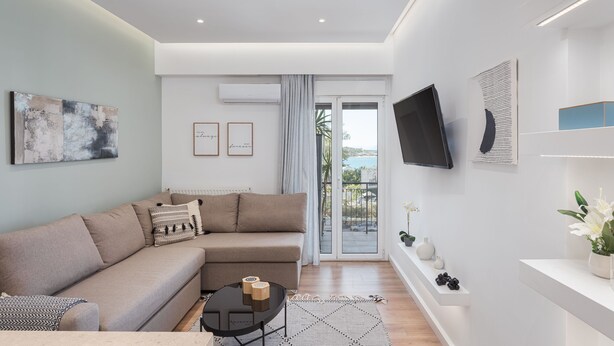 A modern living room with a sofa, a coffee table, and a TV mounted on the wall. There is a sliding glass door leading to a balcony. - Glyfada Cozy Living (Glyfada)