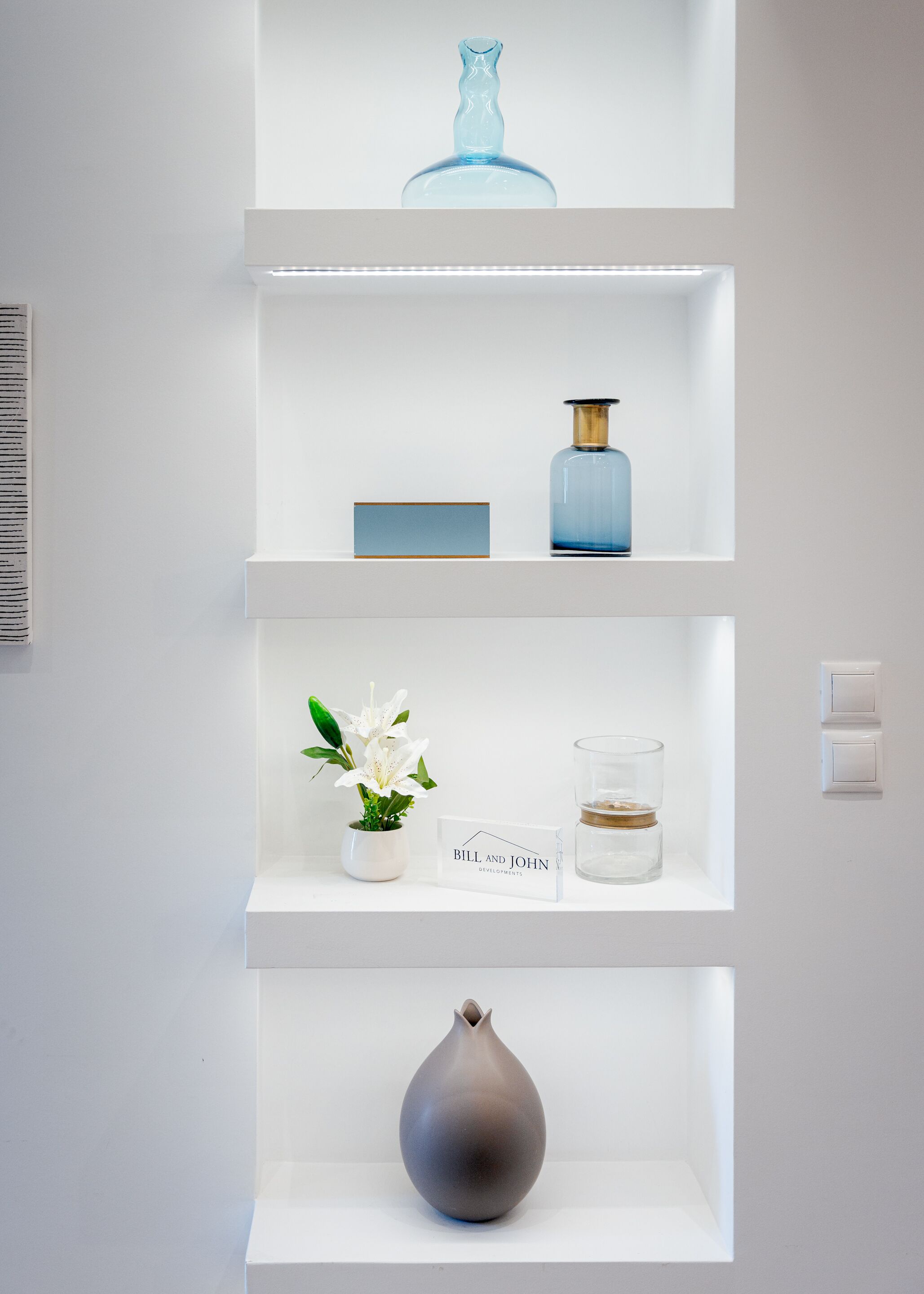 Ein weißes Regal mit einer blauen Vase, einer kleinen blauen Flasche, einer weißen Vase mit weißen Blumen, einem Glas mit Wasser und einem Schild mit der Aufschrift 'Bill and John Developments'.