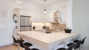 A modern kitchen with white cabinetry, a large island with bar stools, a stainless steel refrigerator, and a microwave. - Glyfada Cozy Living (Glyfada)