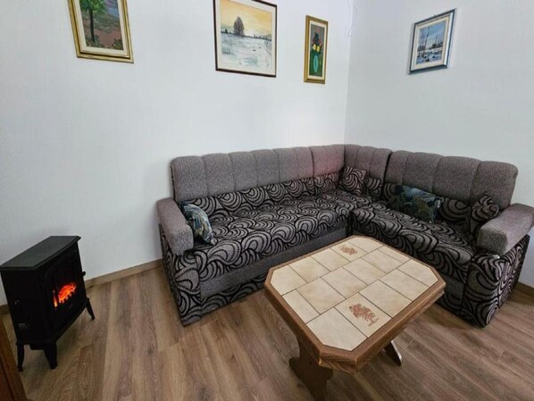 A living room with a grey sectional sofa, a small wooden table, and a black wood stove. - Apartment Gračac (Gracac)