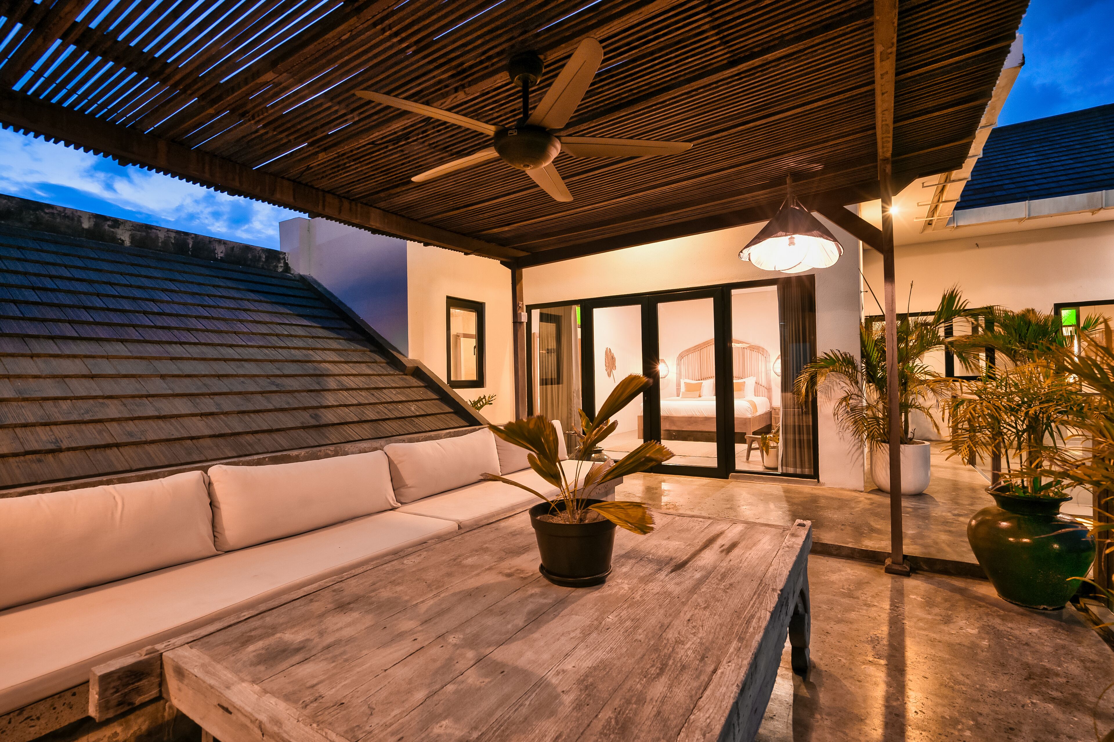 An outdoor patio with a wooden table, a potted plant, a ceiling fan, and a white cushioned bench.