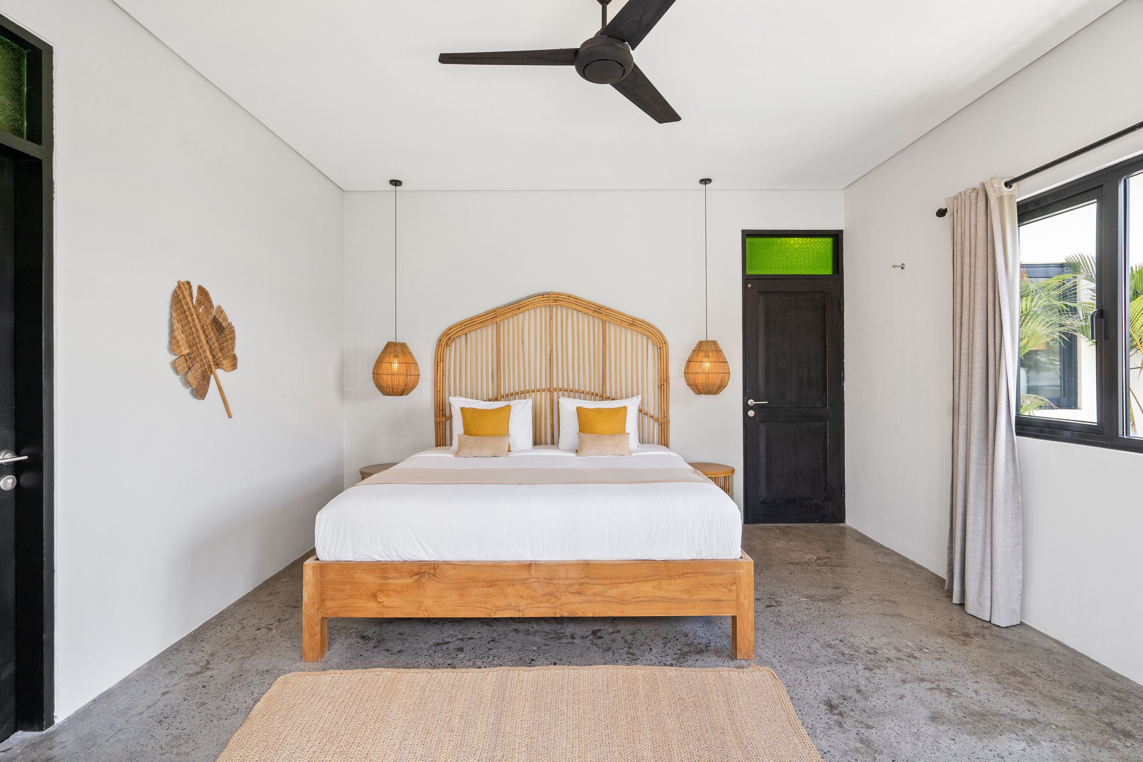 A bedroom with a wooden bed, a ceiling fan, a large window with curtains, and a door with a green light.