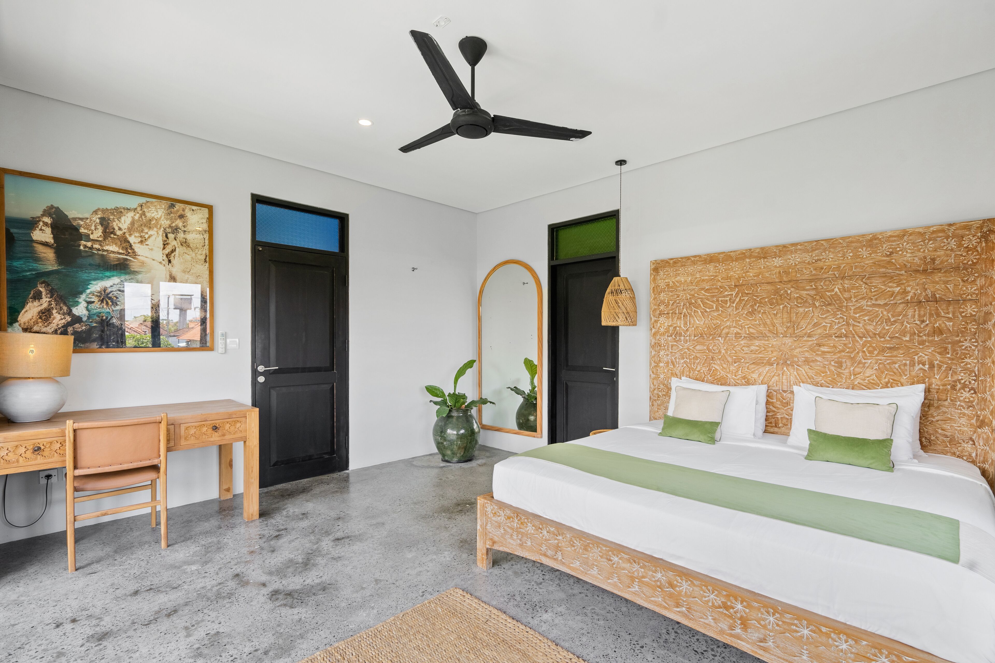 A modern bedroom with a bed, desk, and chair, a ceiling fan, and a large painting of a coastal scene.