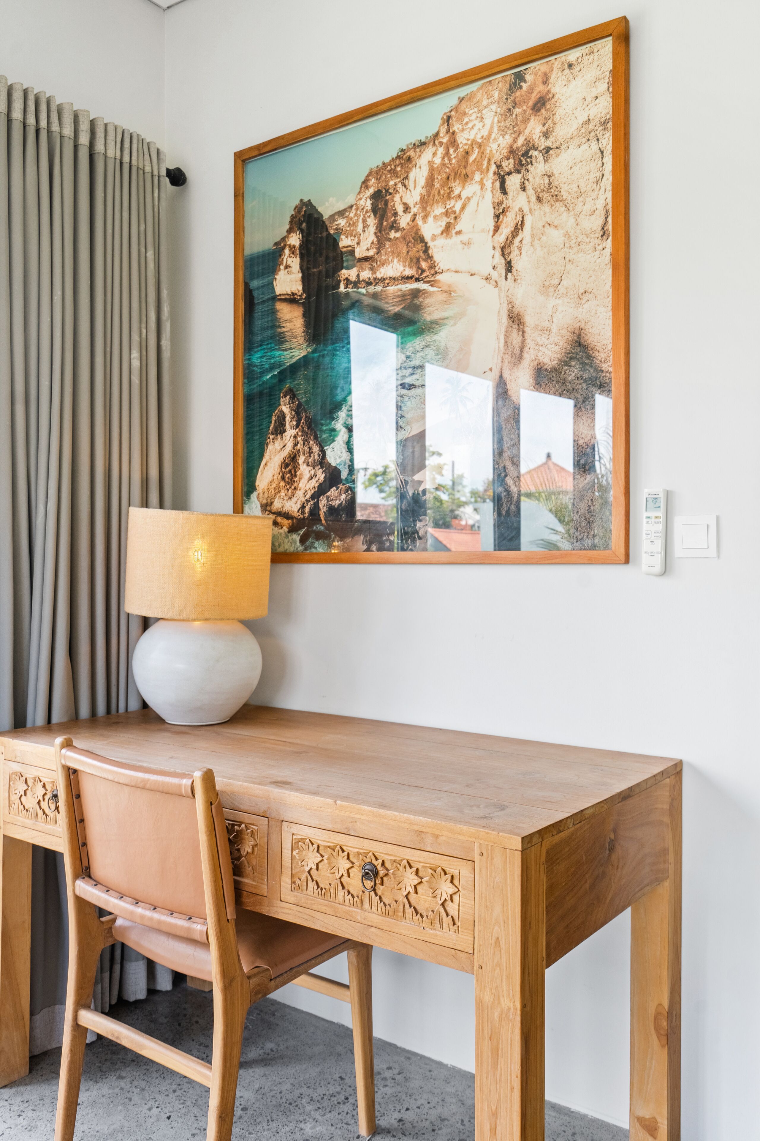 A wooden desk with a lamp, a framed picture of a coastal landscape, and a curtain.