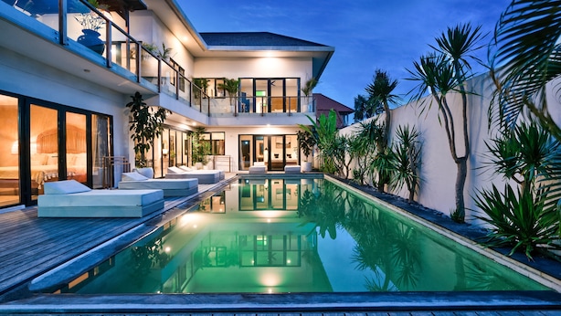Una casa moderna de dos pisos con piscina, mobiliario exterior y plantas tropicales.