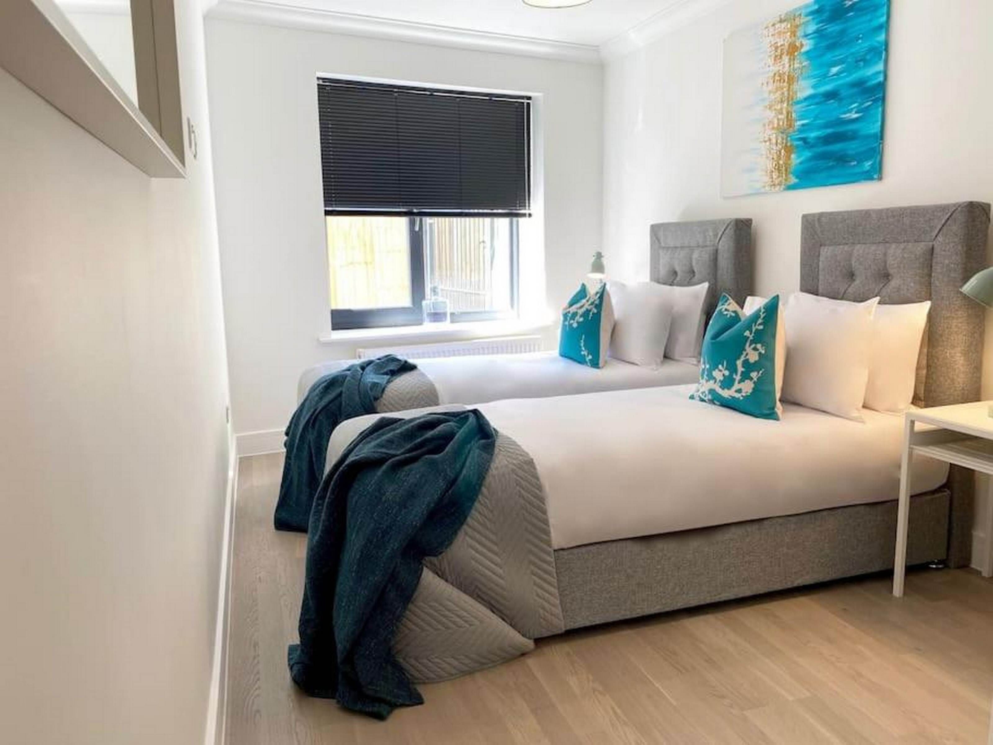 A modern bedroom with a large bed, a bedside table, a window with blinds, and a painting on the wall.