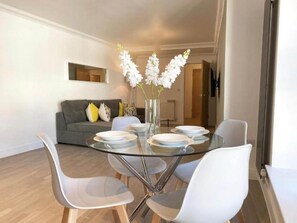 A modern dining area with a glass table surrounded by white chairs, a grey sofa with yellow cushions, and a vase of white flowers.