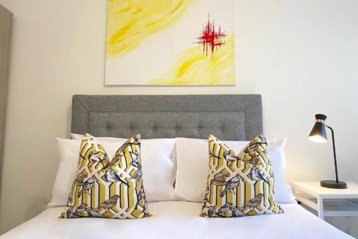 Une chambre d’hôtel avec une tête de lit recouverte d’un tissu gris, du linge de lit blanc et deux coussins décoratifs ornés d’un motif d’oiseaux.