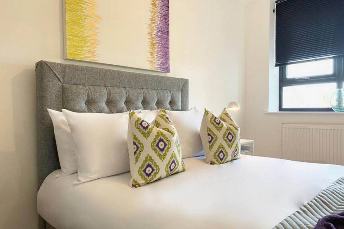 Un lit bien fait, doté d’une tête de lit rembourrée de couleur grise, de draps blancs et de deux coussins décoratifs à motifs géométriques.