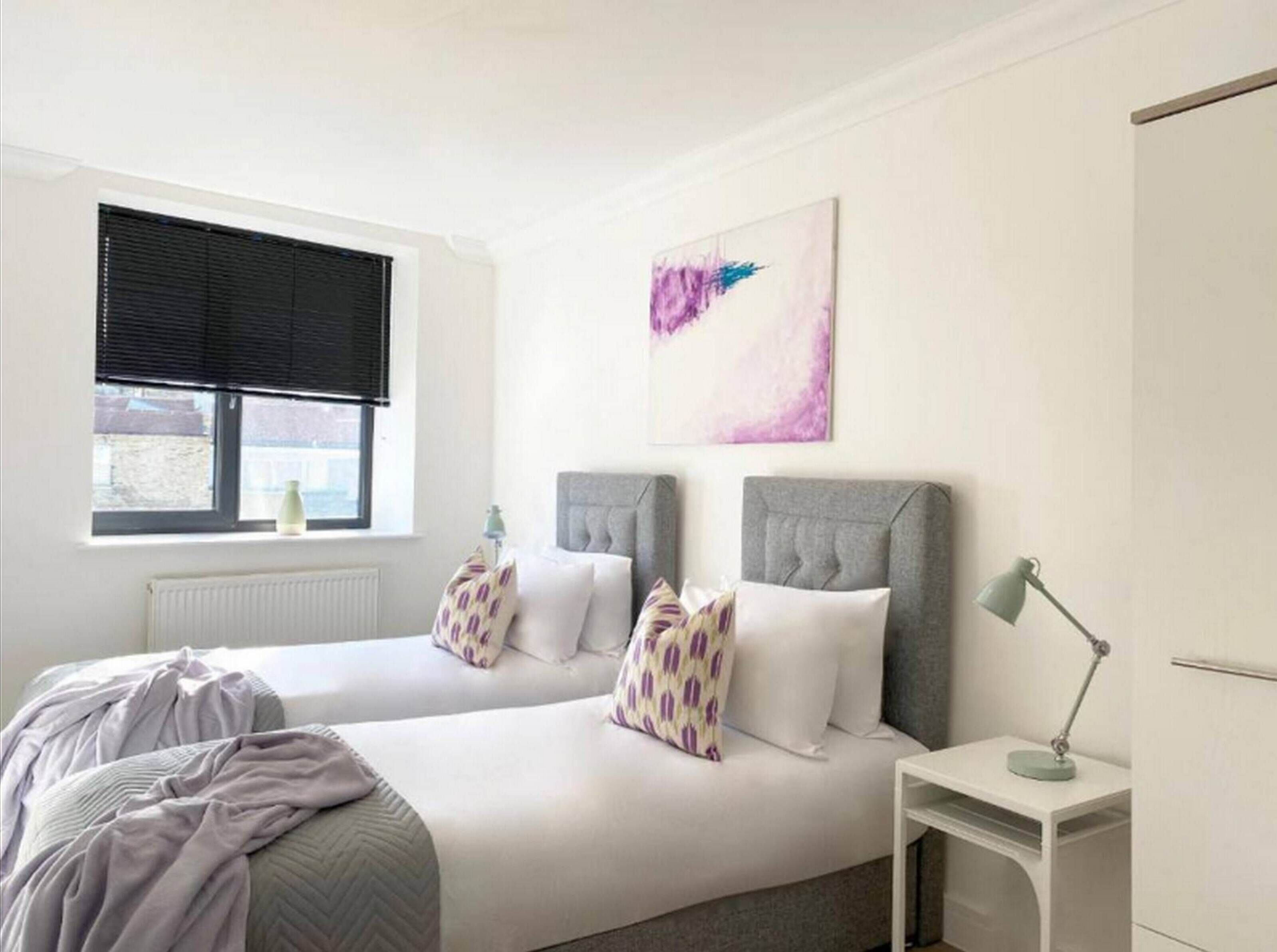 Camera da letto con due letti, una finestra con persiane, un termosifone, un comodino con lampada e un'opera d'arte appesa al muro.
