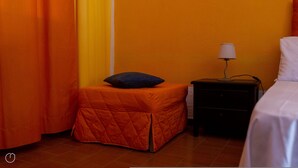 A small, orange-quartered bed with a white pillow, a nightstand with a lamp, and a blue pillow on a matching ottoman. - La Casa di Elisabetta (Budoni)
