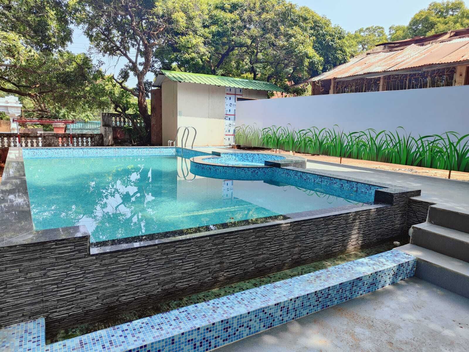 Una piscina con pared de piedra y escalones de azulejos, rodeada de vegetación y un edificio con techo verde.