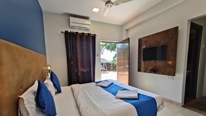 A hotel room with a bed, a ceiling fan, an air conditioning unit, a TV mounted on the wall, and a view of the outside. - Rain Raga Resort (Mahabaleshwar)