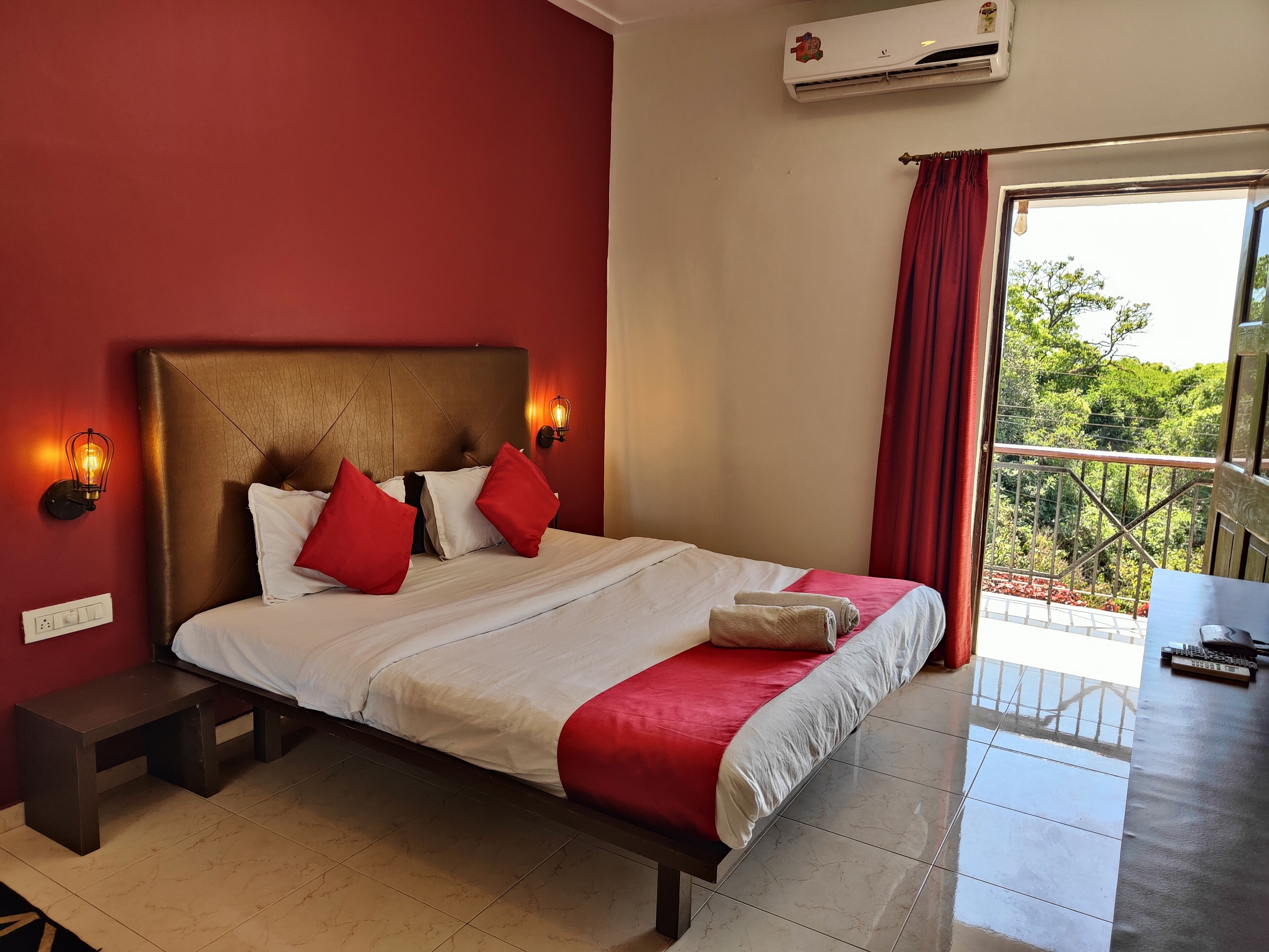 A hotel room with a large bed, red and white bedding, a wooden headboard, a nightstand, a bench, a wall-mounted light, and a view of greenery through a balcony door.
