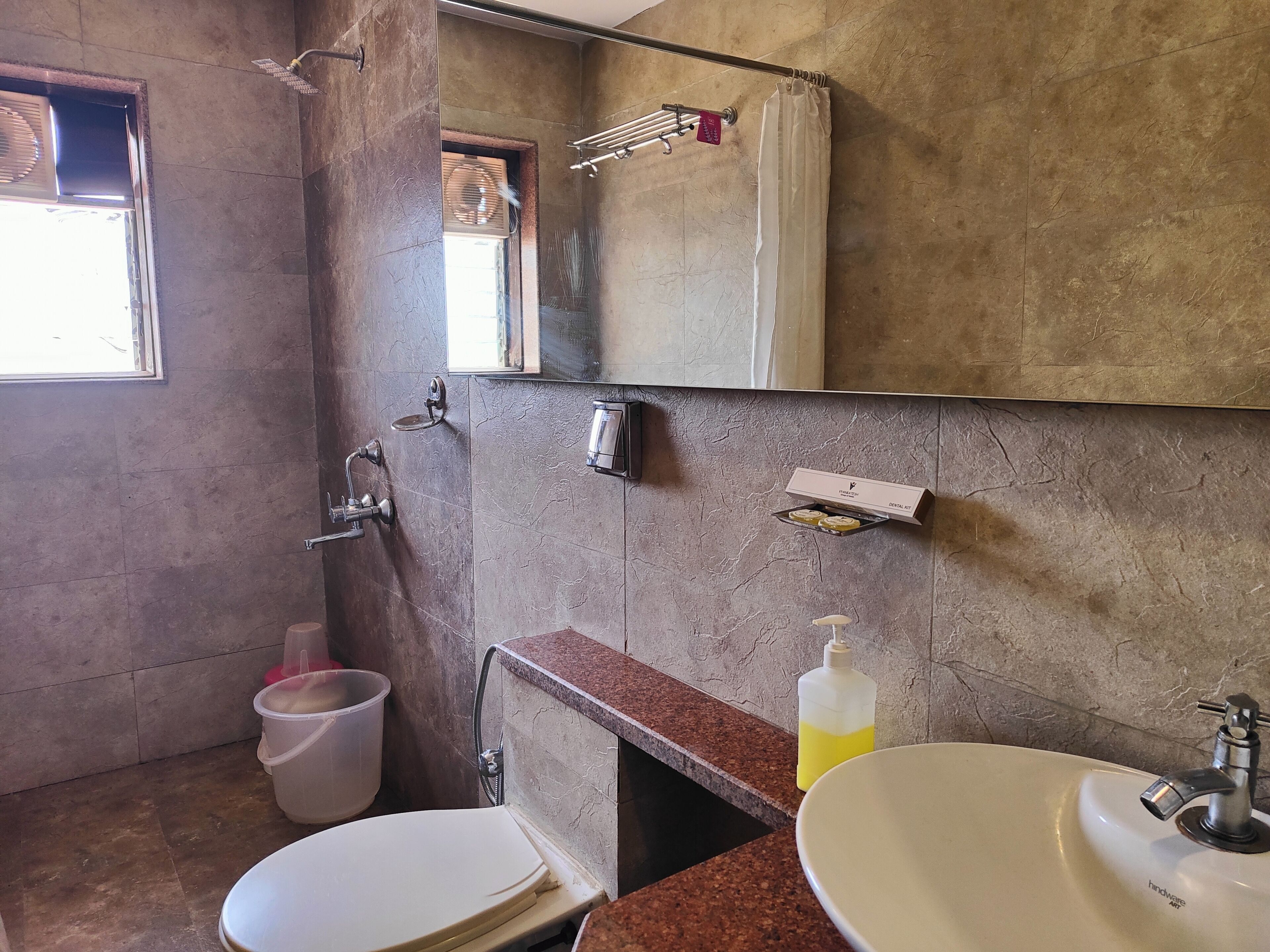 A bathroom with a toilet, sink, and shower. There is a window, a towel rack, and a soap dispenser.