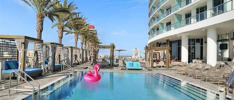 A swimming pool with lounge chairs and a pink flamingo float, surrounded by palm trees and a modern building.