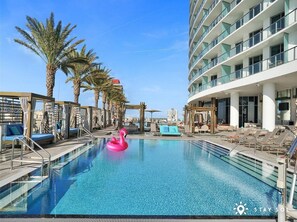 A swimming pool with lounge chairs and a pink flamingo float, surrounded by palm trees and a modern building.