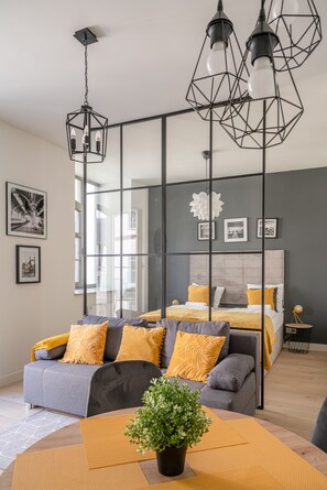 A modern bedroom with a grey and yellow color scheme, featuring a bed with a grey headboard, a grey sofa with yellow cushions, and a large window providing natural light. - RentPlanet - Młyn Maria (Wroclaw)