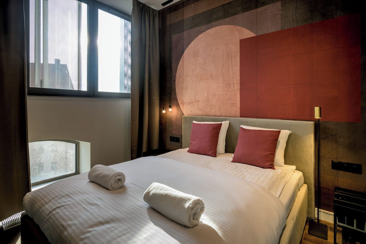 Quarto de hotel com cama grande, duas almofadas, janela com cortinas e parede com um desenho circular.