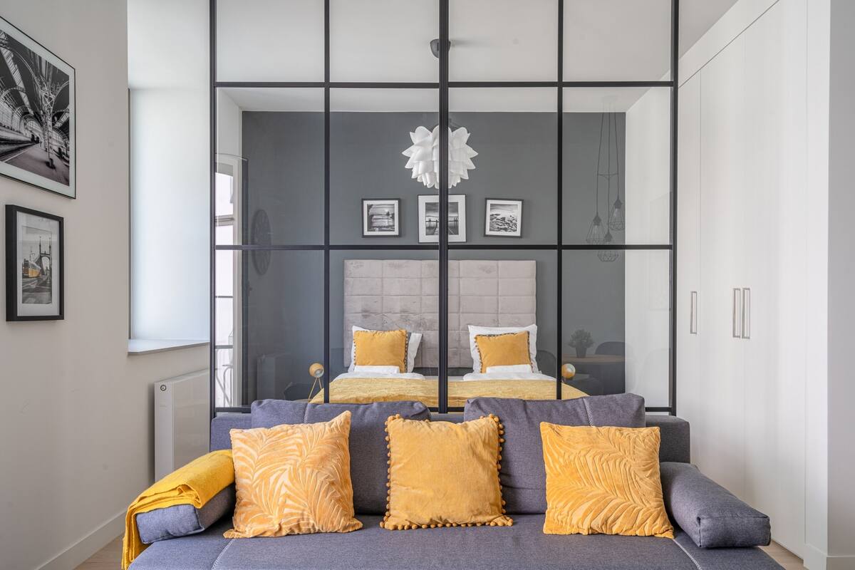 Quarto moderno com sofá cinza, almofadas amarelas e um espelho grande refletindo o ambiente.