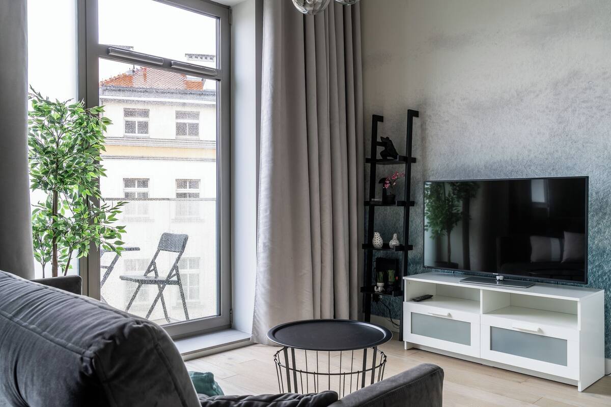 Uma sala de estar moderna com uma janela grande, uma TV de tela plana, um rack branco, um sofá cinza e uma mesa redonda pequena.