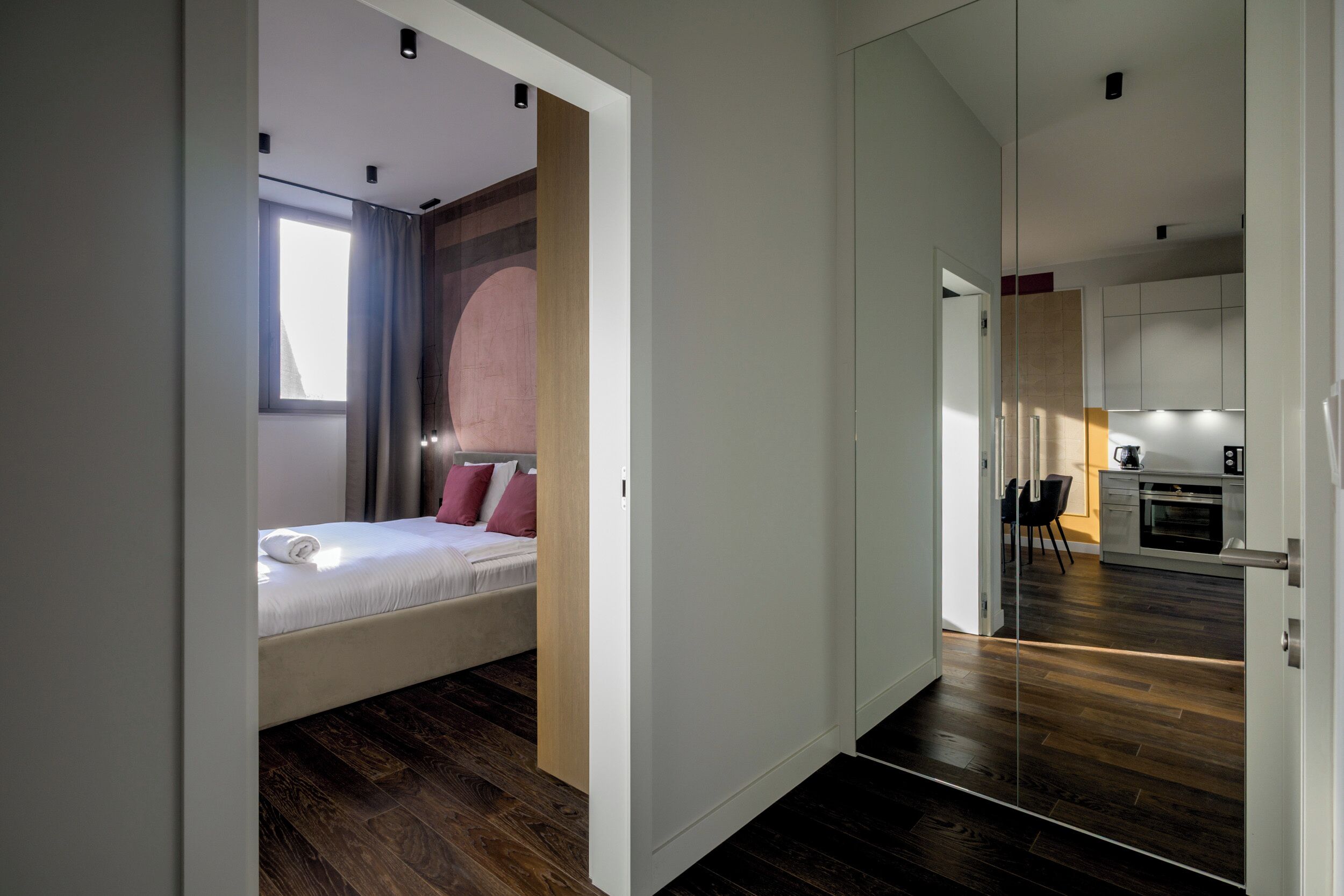 A modern hotel room with a bed, a small seating area, and a view of the kitchen.