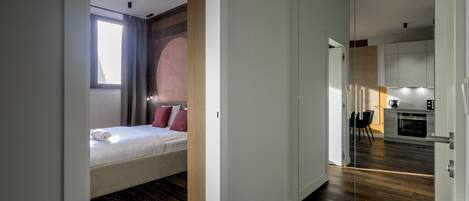 A modern hotel room with a bed, a small seating area, and a view of the kitchen.