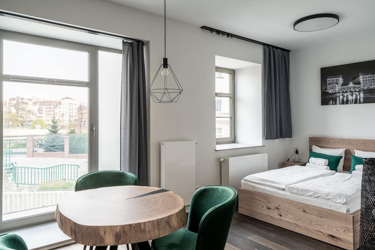 Quarto de hotel moderno com cama de madeira, cadeiras verdes, mesa redonda de madeira e grandes janelas com cortinas.