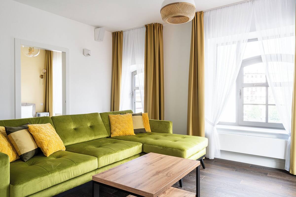 Um sofá modular verde com almofadas amarelas, uma mesa de centro de madeira e uma janela com cortinas leves.