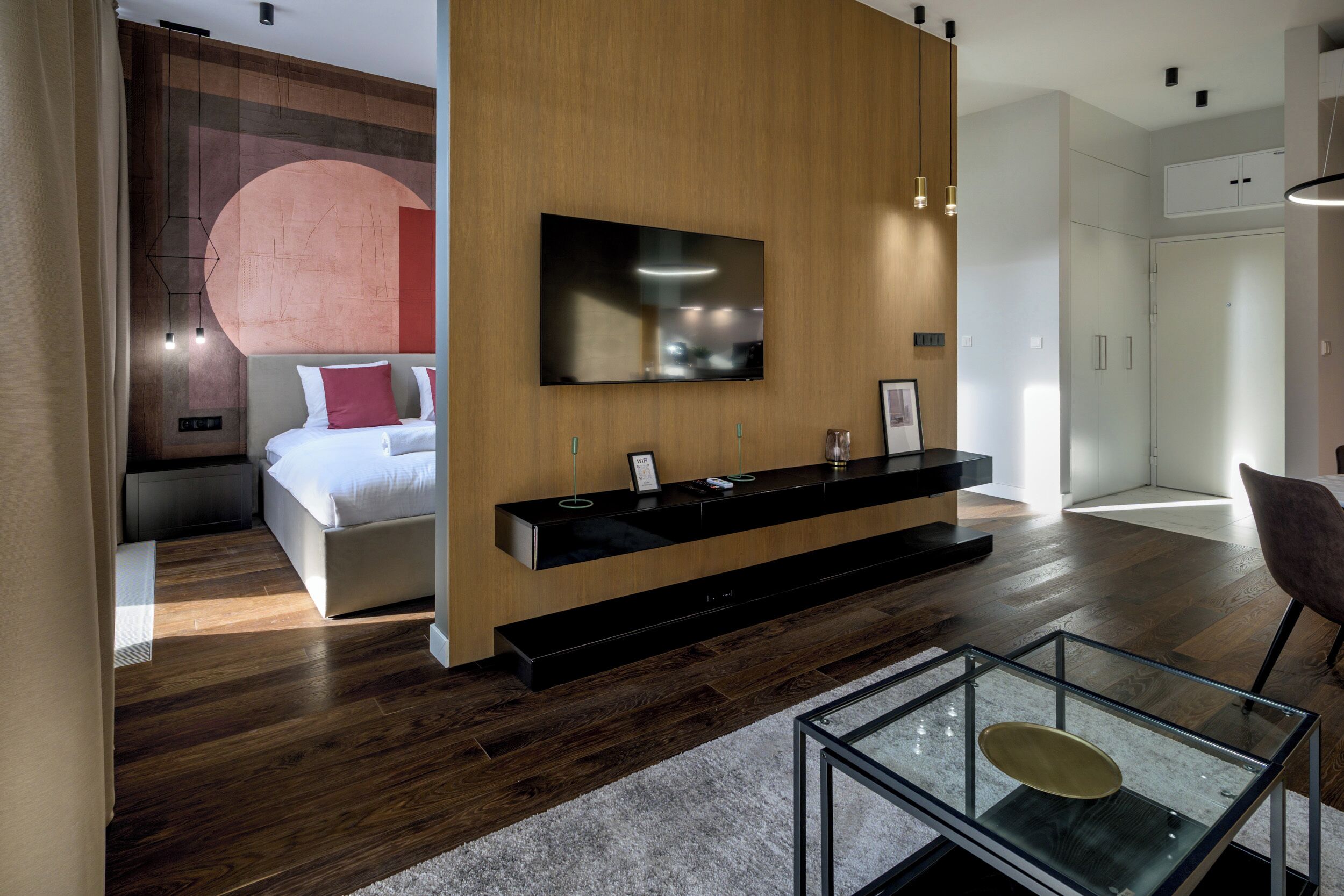 A modern hotel room with a wooden accent wall, a large flat-screen TV, a built-in black console table, a glass coffee table, and a large bed with red and white bedding.