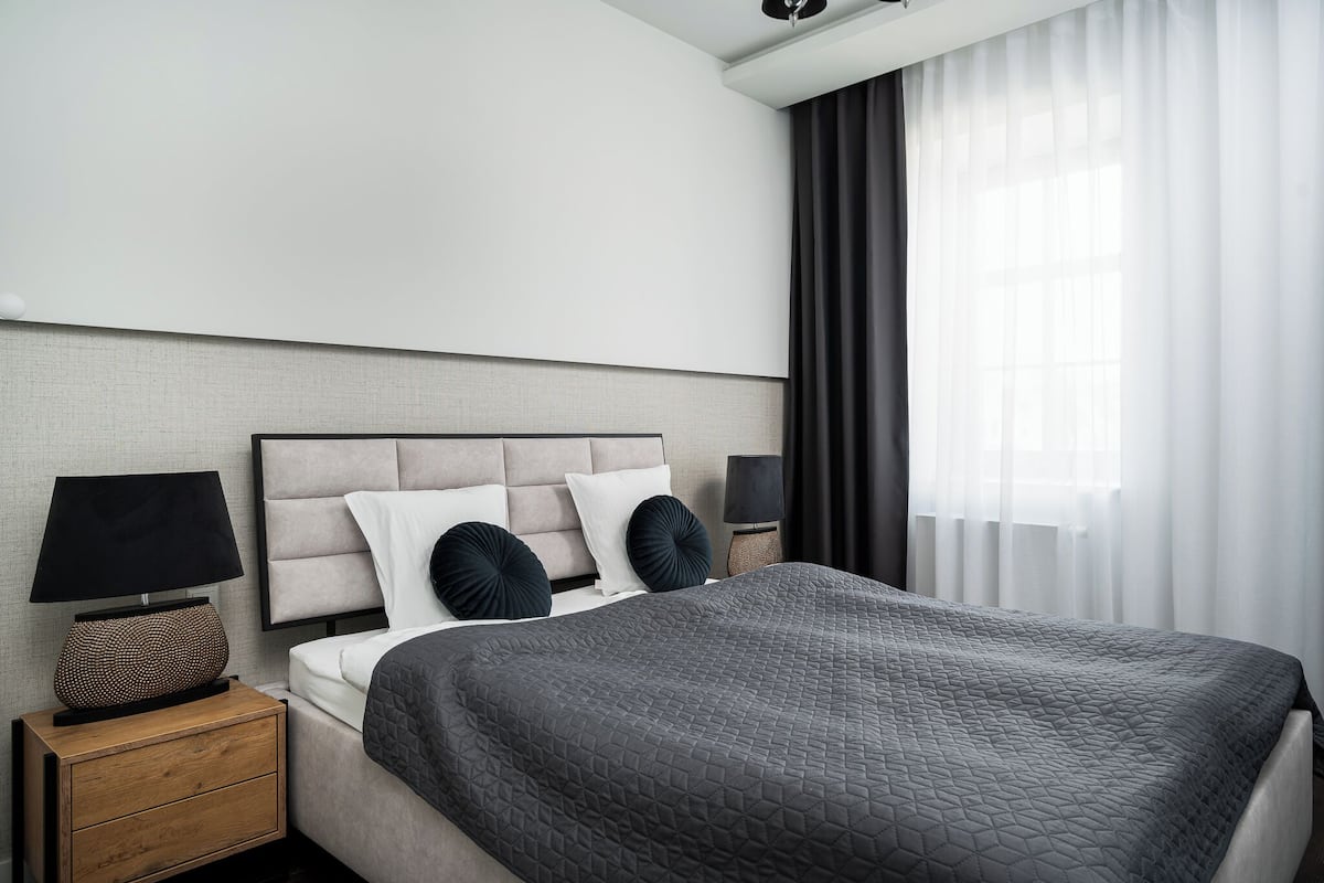 Quarto de hotel moderno com uma cama grande, uma mesa de cabeceira com abajur e vista para a cidade através de cortinas leves.