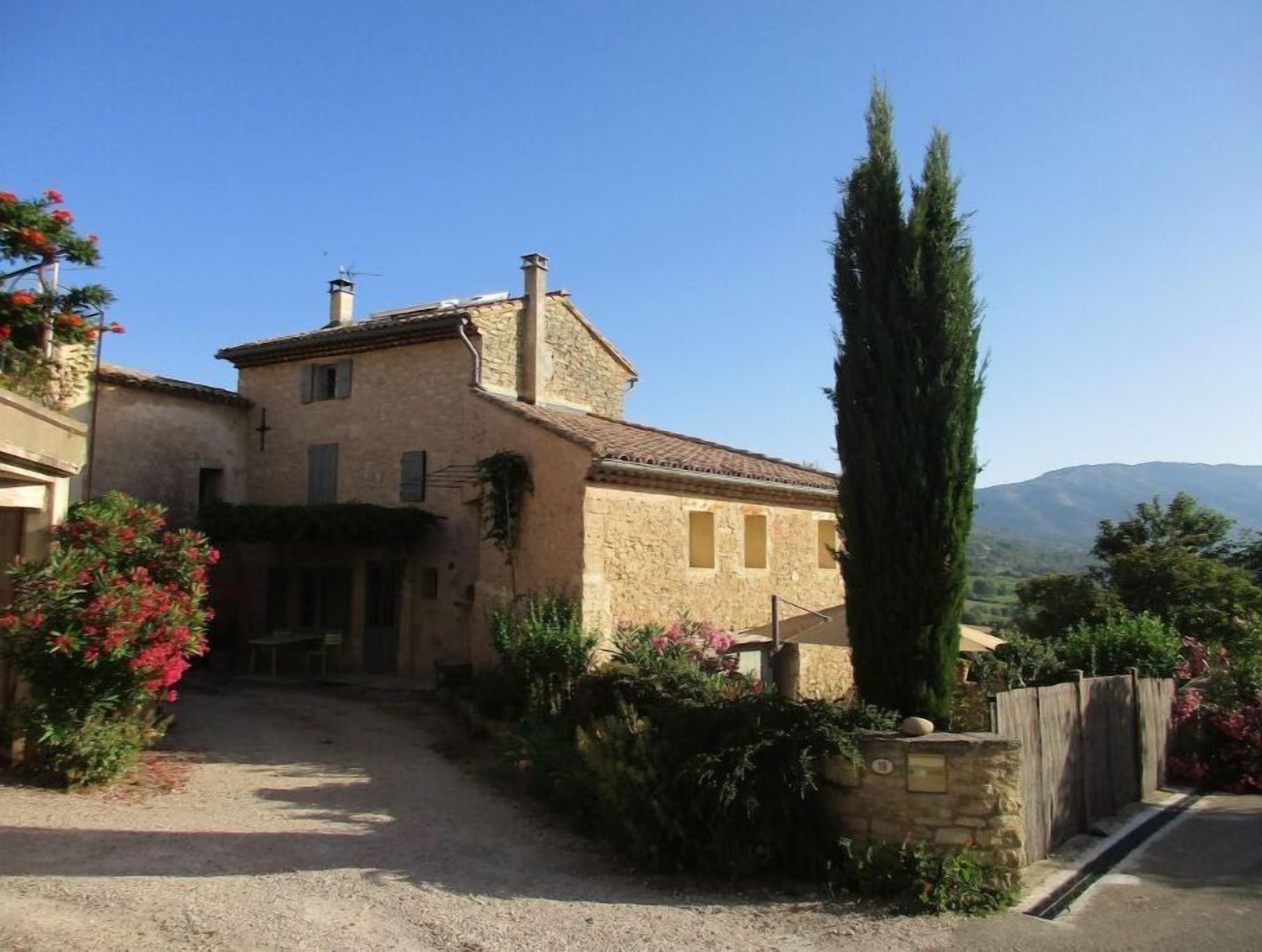 A stone house with a tiled roof, a chimney, and a garden with shrubs and flowers.