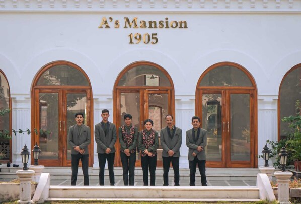 Un groupe d’hommes se tient devant un bâtiment sur lequel on peut lire l’inscription « Manoir A, 1905 », et qui est doté de grandes fenêtres en arc de cercle.