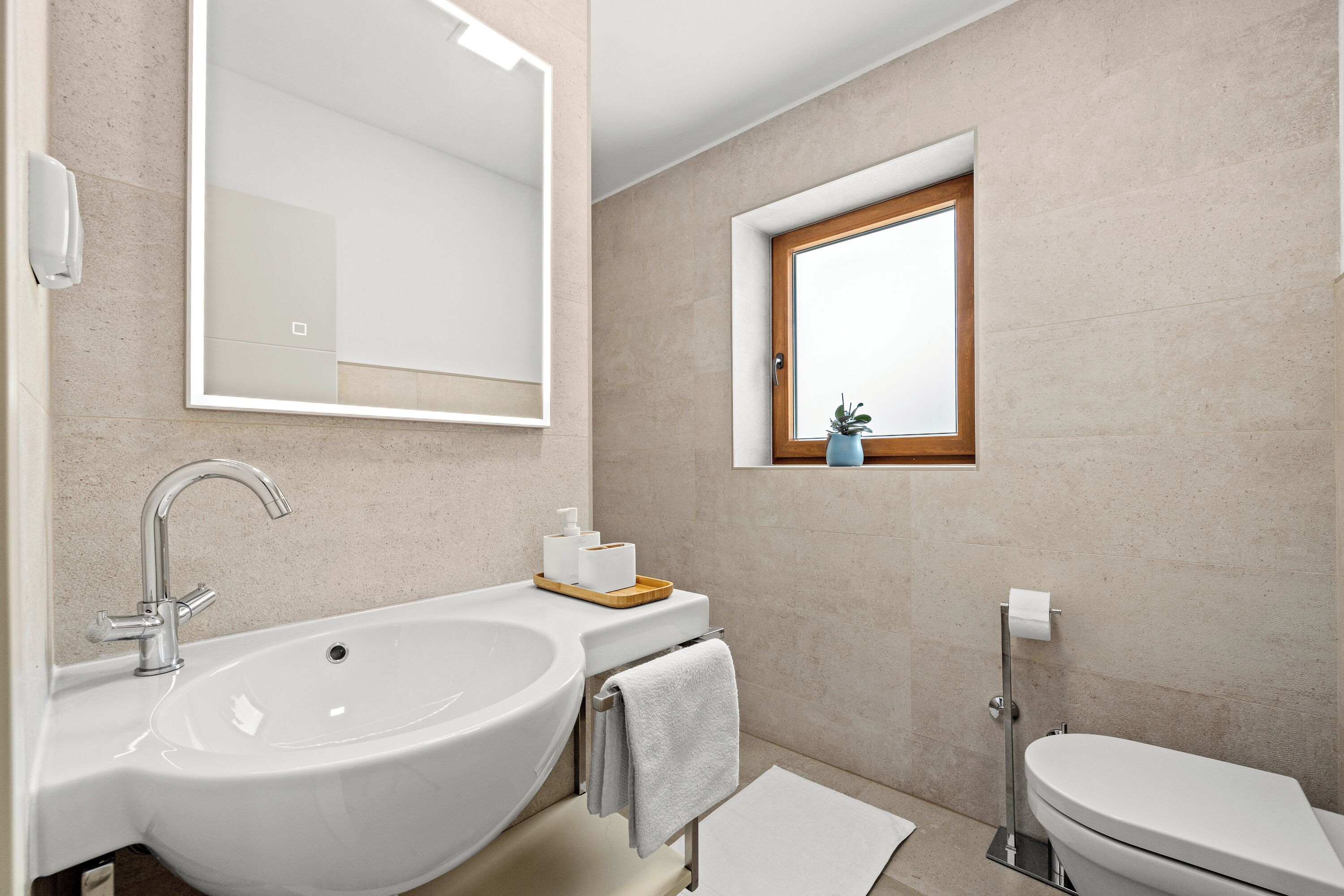 A modern bathroom with a large sink, a toilet, and a small window with a plant.