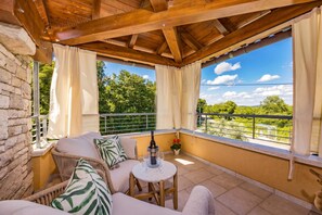 Ein Balkon mit Holzboden, einer Steinwand, einer gepolsterten Sitzecke, einem kleinen Tisch mit Flasche und Gläsern sowie Blick ins Grüne.