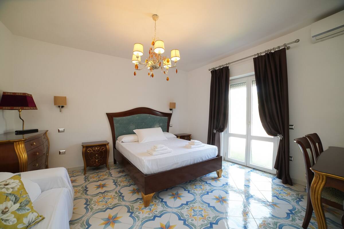 A hotel room with a large bed, a chandelier, a dining table, and a view of the outdoors.