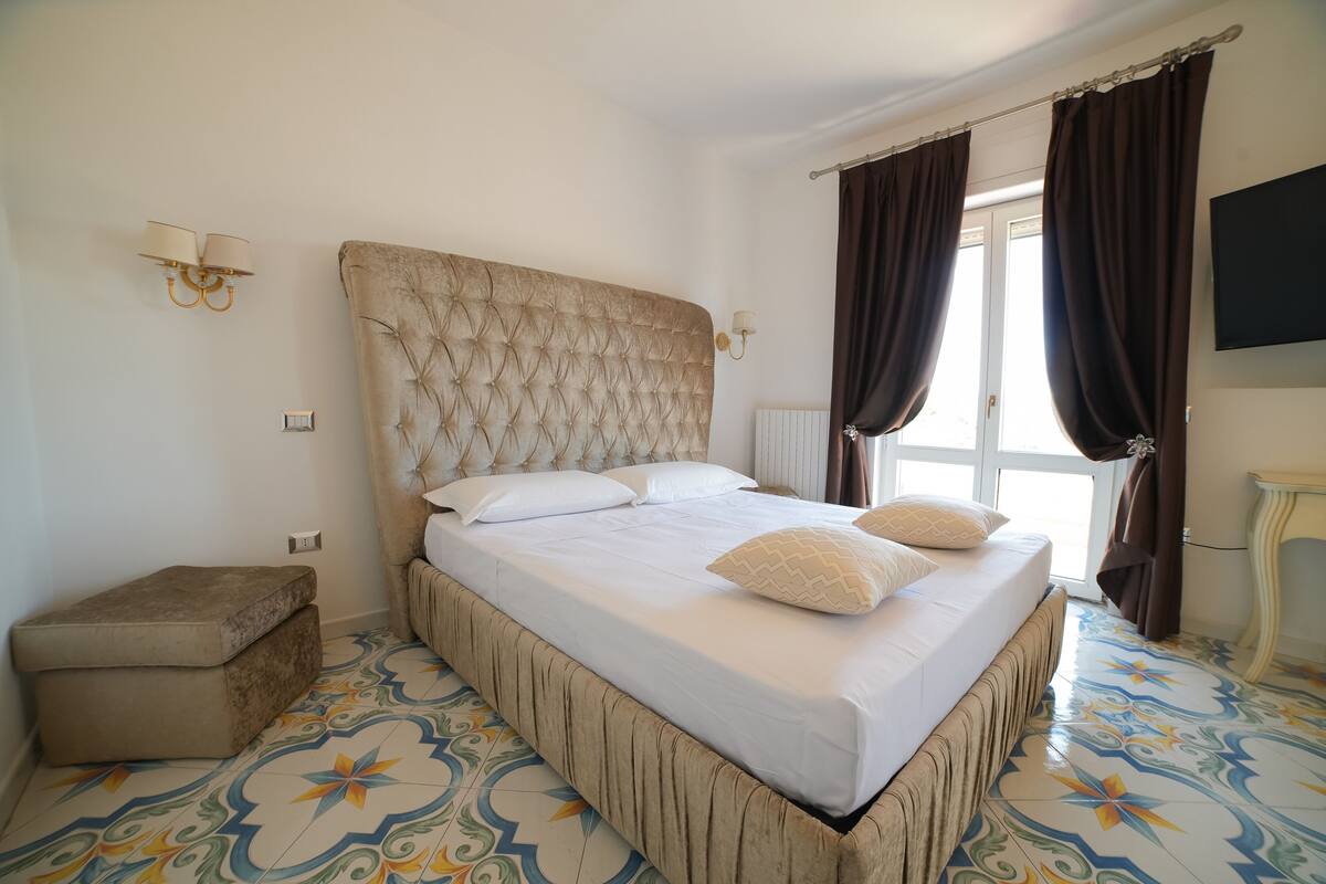 A bedroom with a tufted headboard, a bed with white linens, a bench, patterned floor tiles, and a television on the wall.