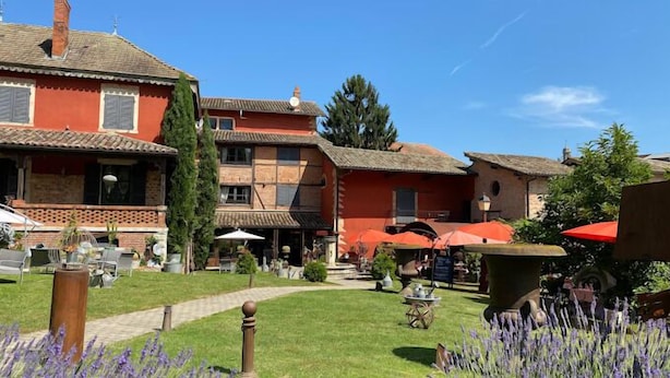 A courtyard with a building featuring a tiled roof and a red facade. There are outdoor tables and chairs, and a pathway leading to the building. - Les Jardins de la Tour (Chatillon-sur-Chalaronne)