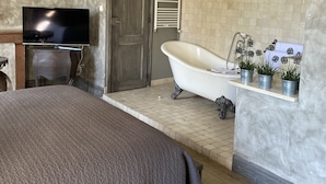 A bedroom with a large bed, a television, a clawfoot bathtub, and a shelf with potted plants. - Les Jardins de la Tour (Chatillon-sur-Chalaronne)