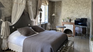 A bedroom with a bed, curtains, a TV, a fireplace, and a small table. - Les Jardins de la Tour (Chatillon-sur-Chalaronne)
