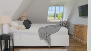 Et soveværelse med skrå vægge, en seng med hvide sengetæpper, et gråt pled, en sengetavlelampe, et træskab og et vindue med udsigt til grønne områder.