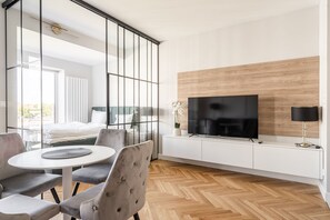 Une chambre d’hôtel moderne dotée d’une grande télévision, d’un coin repas avec une table ronde et des chaises, et d’un lit offrant une vue sur l’extérieur.