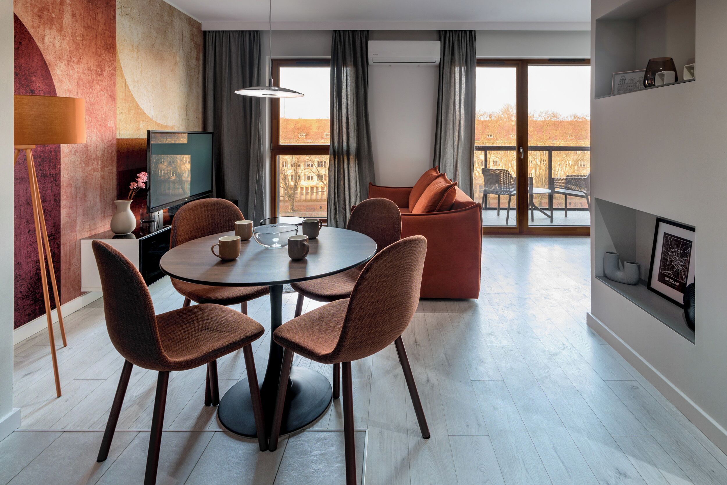 A modern living room with a round dining table, four chairs, a sofa, and a balcony with a view of buildings.