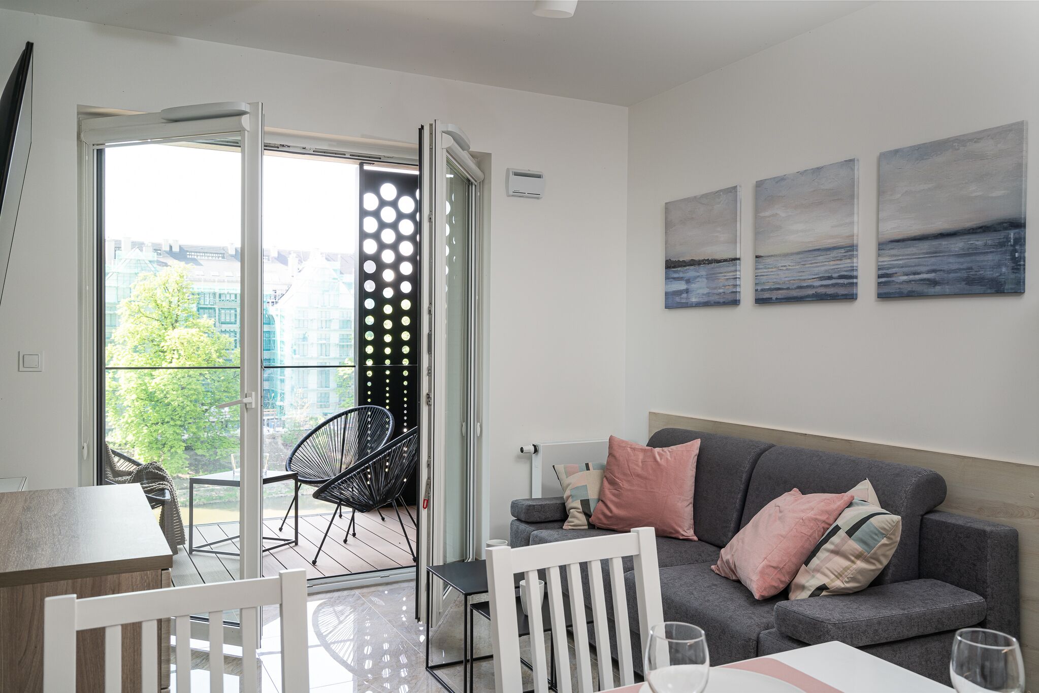 A modern living room with a grey sofa, white chairs, and a glass table. A sliding glass door leads to an outdoor area with chairs.