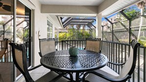Un patio techado al aire libre con una mesa redonda y sillas, un adorno verde y vistas a una piscina y follaje.