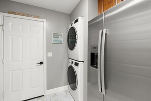 Un lavadero con una puerta blanca, lavadora y secadora empotradas en la pared, y un refrigerador de acero inoxidable.