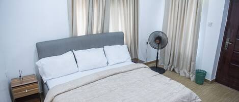 Un lit bien fait, avec une tête de lit grise, une table de chevet avec une lampe et un ventilateur sur pied.