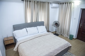 Un lit bien fait, avec une tête de lit grise, une table de chevet avec une lampe et un ventilateur sur pied.
